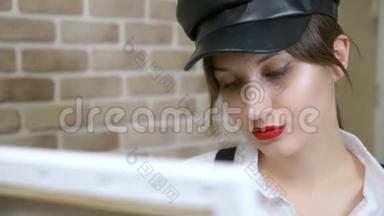 戴皮帽的女艺术家在画布上靠砖墙画一幅画